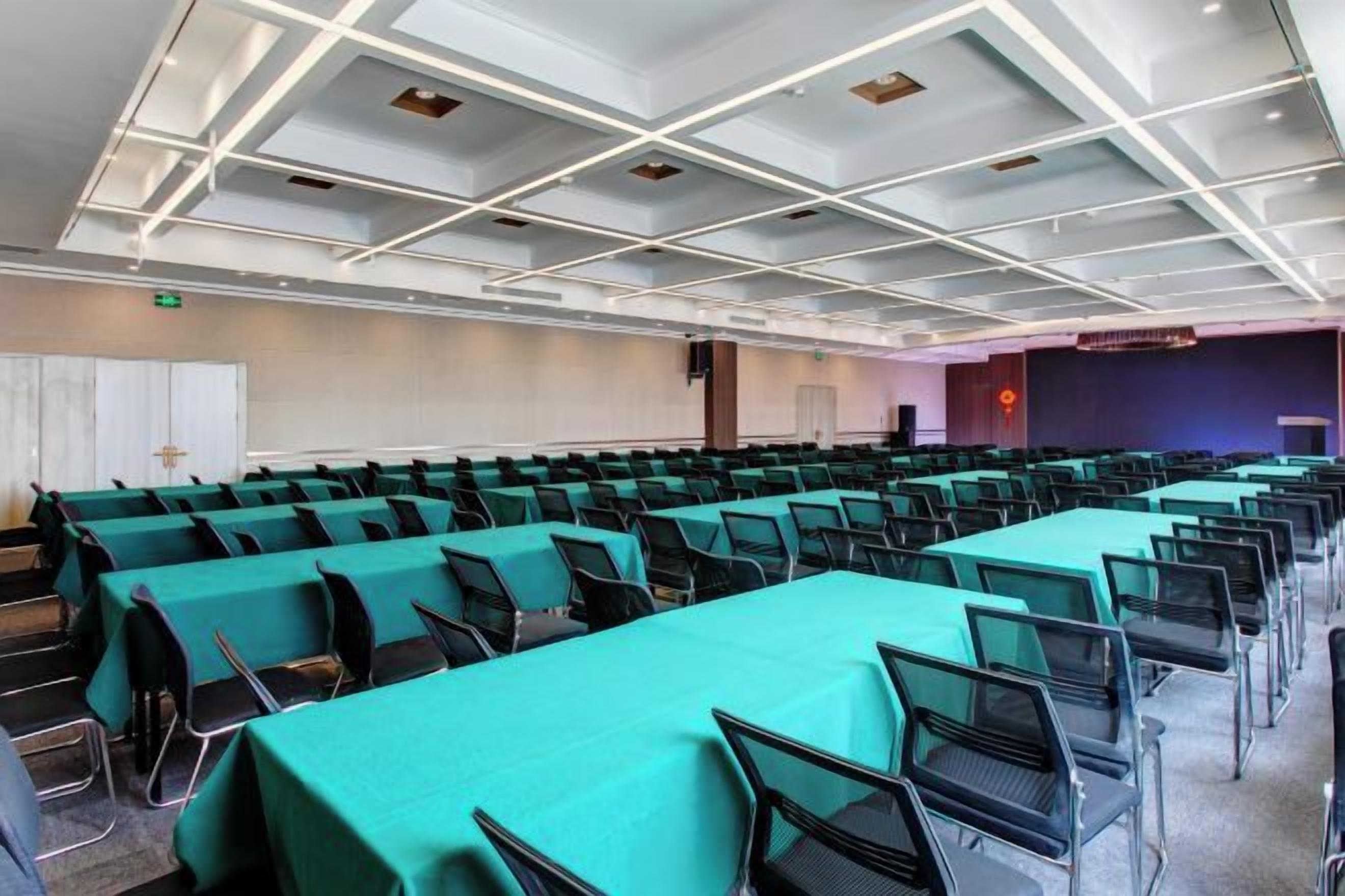 A conference room with rows of tables and chairs, a stage, and a large screen.
