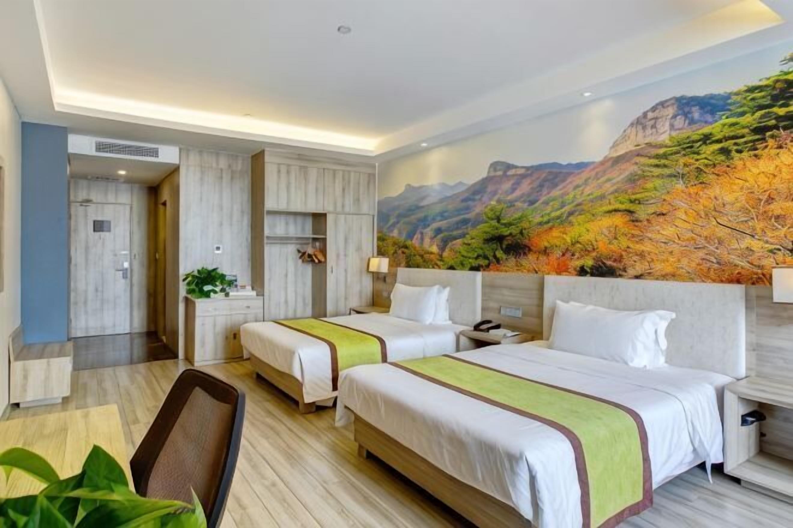 A hotel room with two beds, a desk, a chair, a painting of a mountain landscape, and a plant.