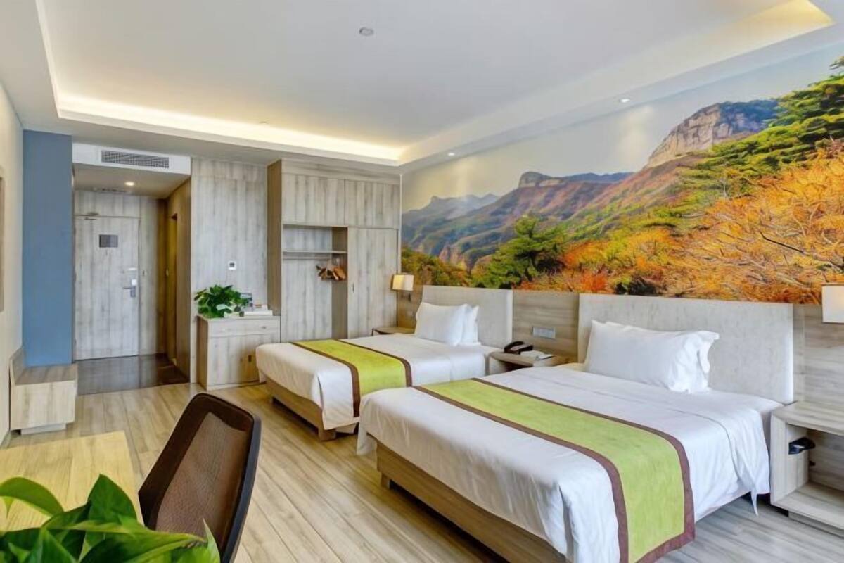 Ein Hotelzimmer mit zwei Betten, einem Schreibtisch, einem Stuhl, einem Gemälde einer Berglandschaft und einer Pflanze.