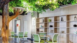 A modern indoor space with a central table surrounded by green chairs, a wooden bookshelf with various decorative items, and a large artificial tree. - Yuncheng Juhetai Hotel (Yanhu South Wind Square) (Yuncheng)