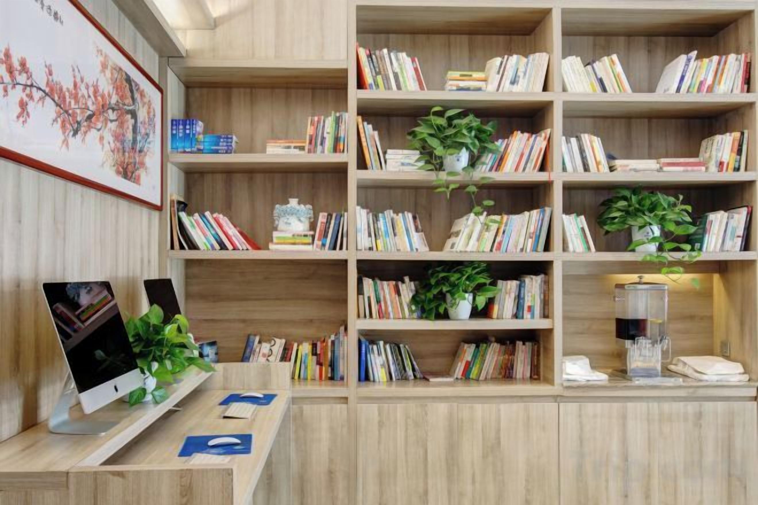 A wooden bookshelf with books and plants, a desk with a computer and a plant, and a framed picture on the wall.