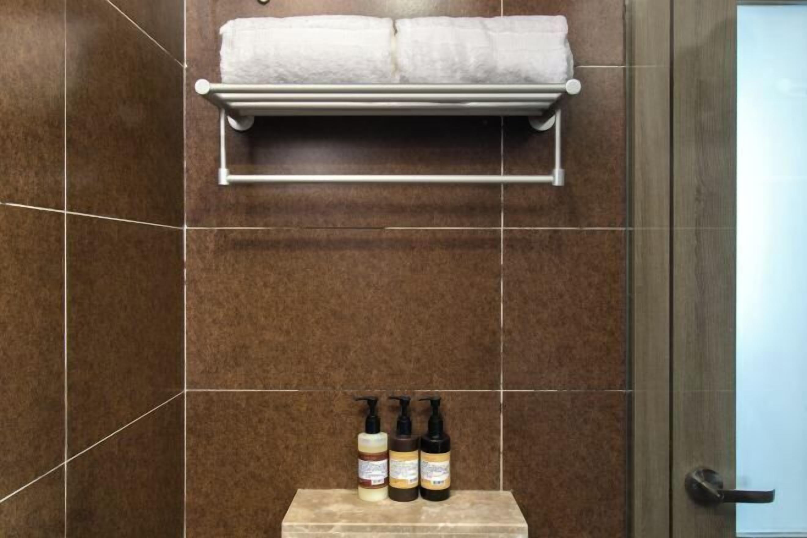 A bathroom with brown tiles, a towel rack with a white towel, and three bottles of toiletries.