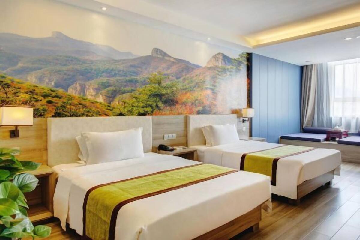 Ein Hotelzimmer mit zwei Betten, einem großen Wandgemälde einer Berglandschaft und einem Fenster mit Vorhängen.