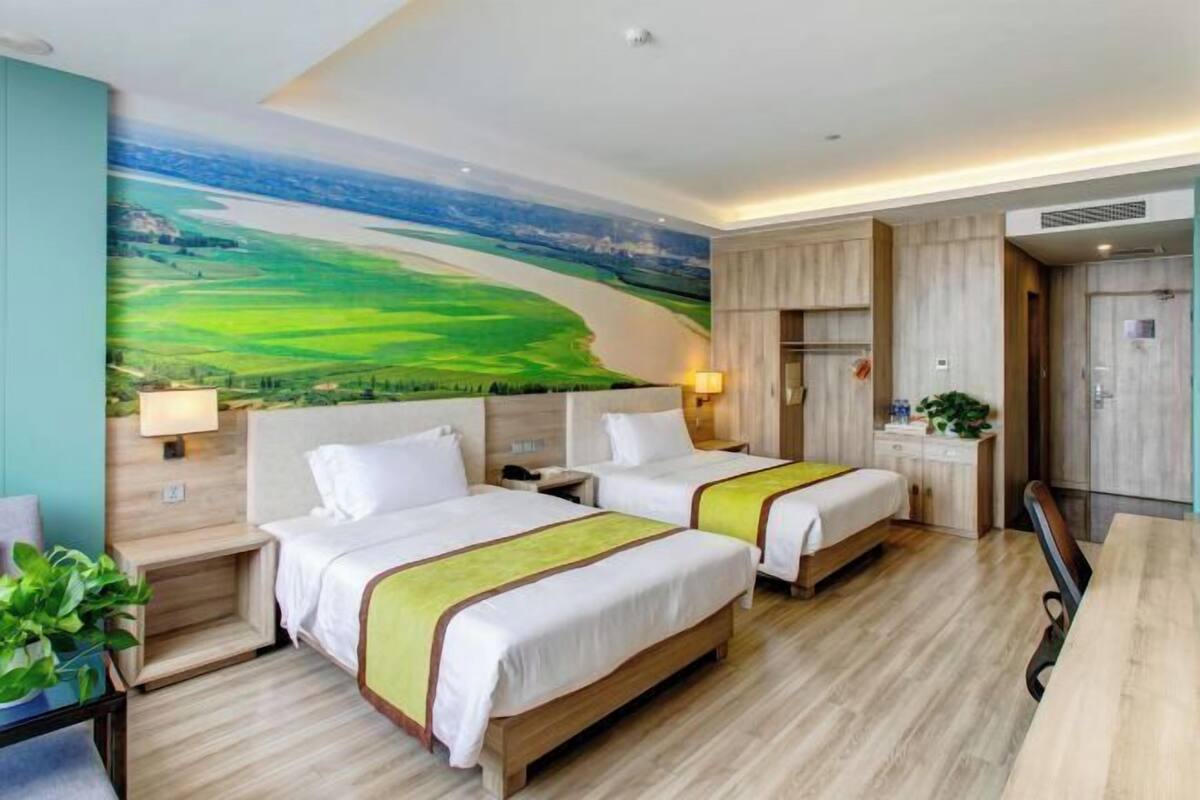 Ein Hotelzimmer mit zwei Betten, einem Schreibtisch, einem Stuhl, einer Lampe, einer Pflanze und einer großen Wandmalerei mit Landschaftsmotiv.