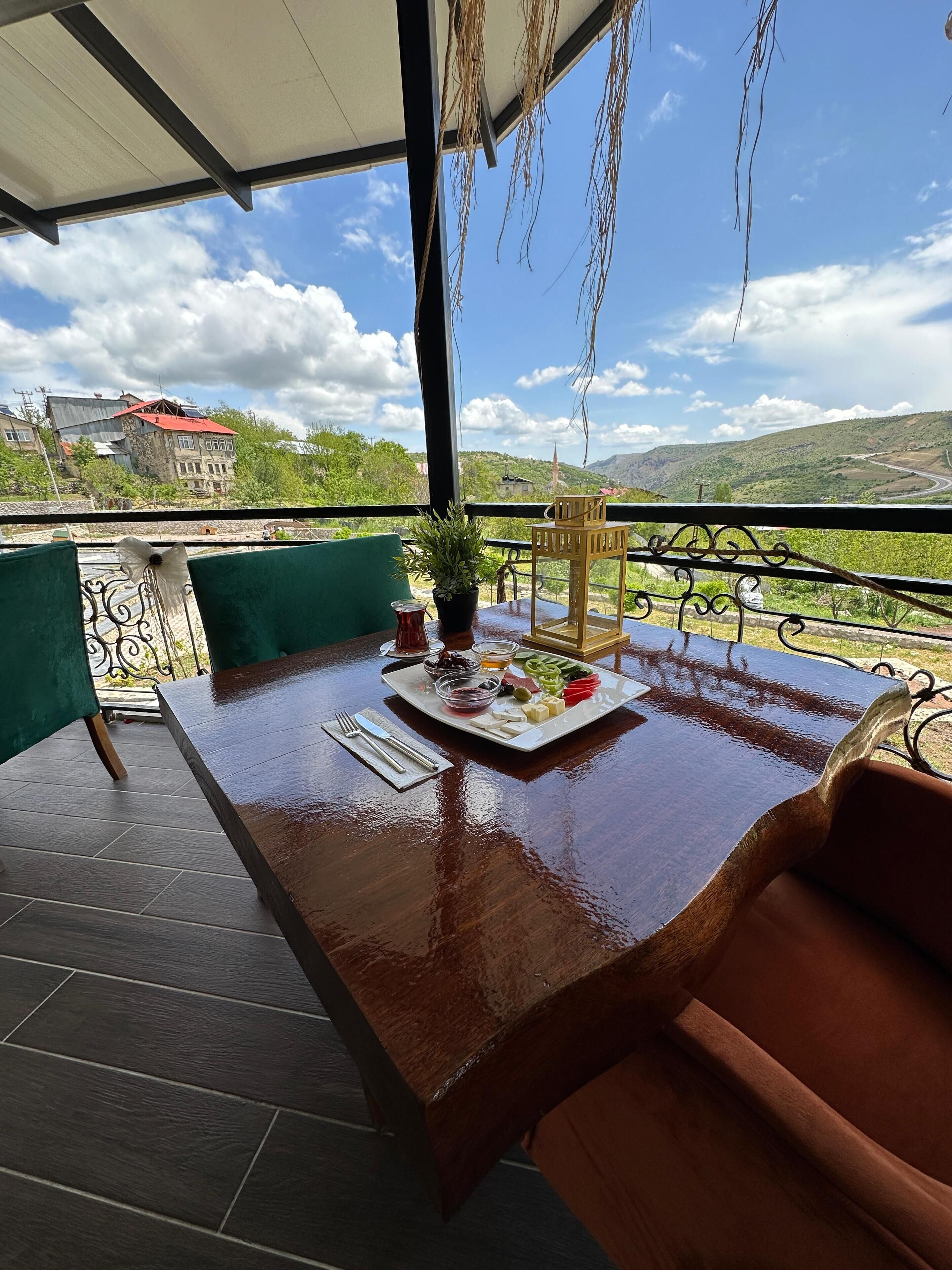 Une table en bois, sur laquelle se trouve une assiette de nourriture et un pot de fleurs, est placée sur un balcon d’où l’on peut admirer un paysage montagneux.