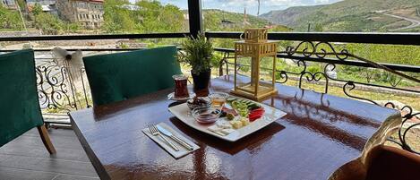 Une table en bois, sur laquelle se trouve une assiette de nourriture et un pot de fleurs, est placée sur un balcon d’où l’on peut admirer un paysage montagneux.