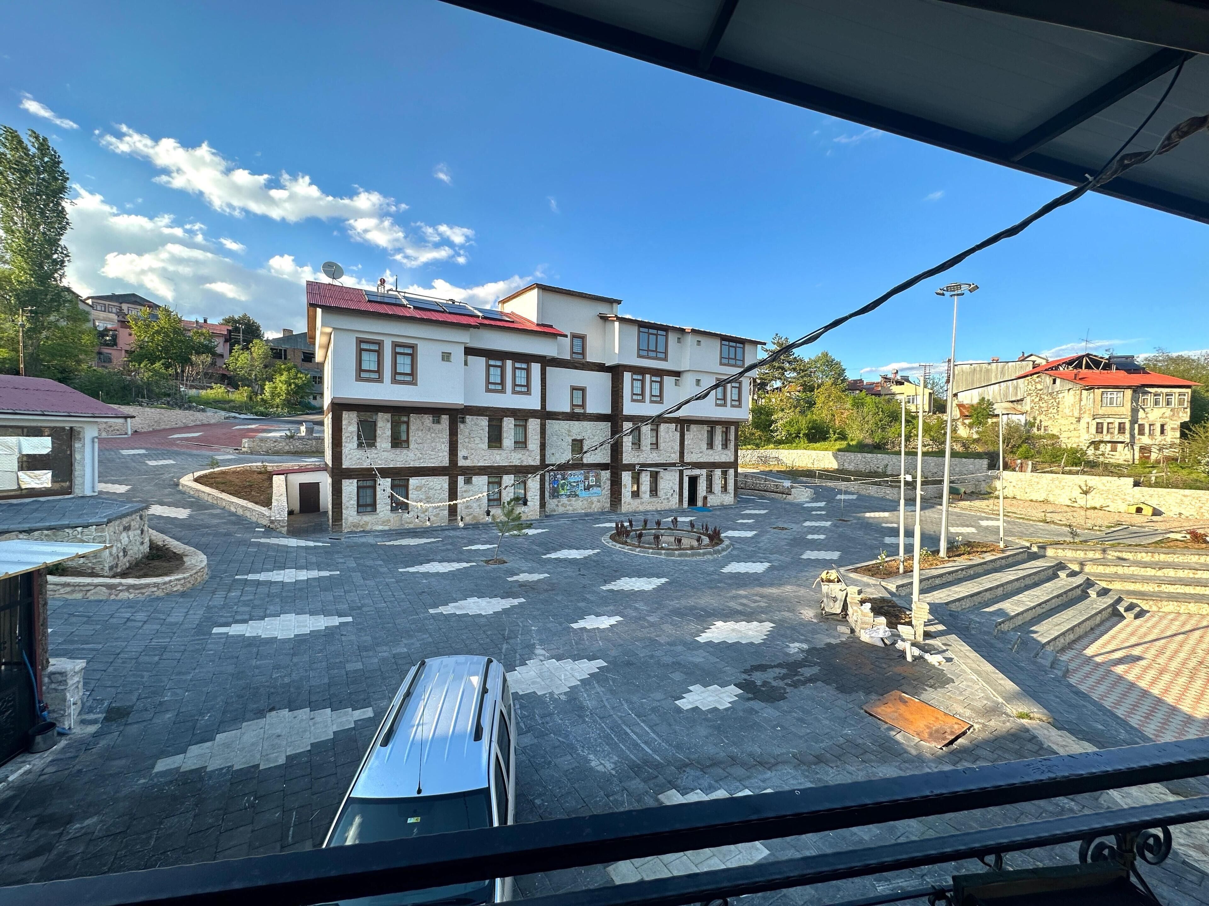 A multi-story building with a mix of stone and wood facade, a central courtyard with patterned paving, and a red-roofed building in the background.