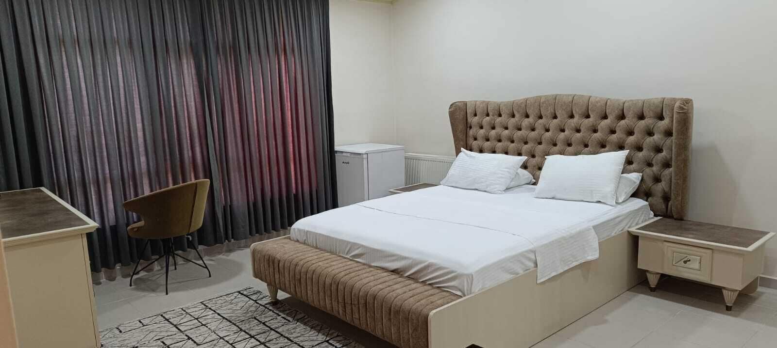 A bedroom with a tufted headboard, a bed with white linens, a bedside table, a chair, and a radiator.