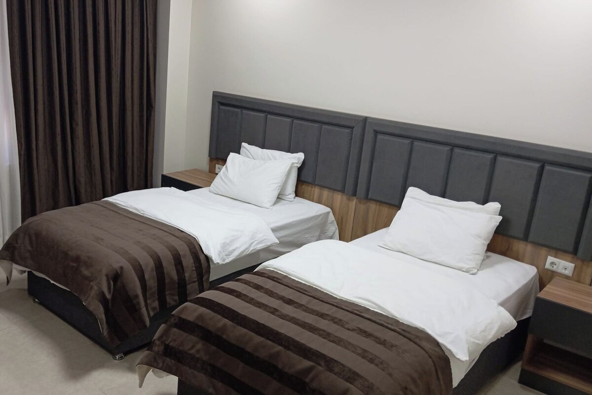 A hotel room with two beds, each with white bedding and brown striped blankets. The room has a grey headboard and a wooden nightstand with a lamp.