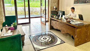 A reception desk with a computer, a framed picture, and a small plant. There is a green chair and a table with a vase and a notebook. - Kaşkaloğlu Hotel (Arapgir)