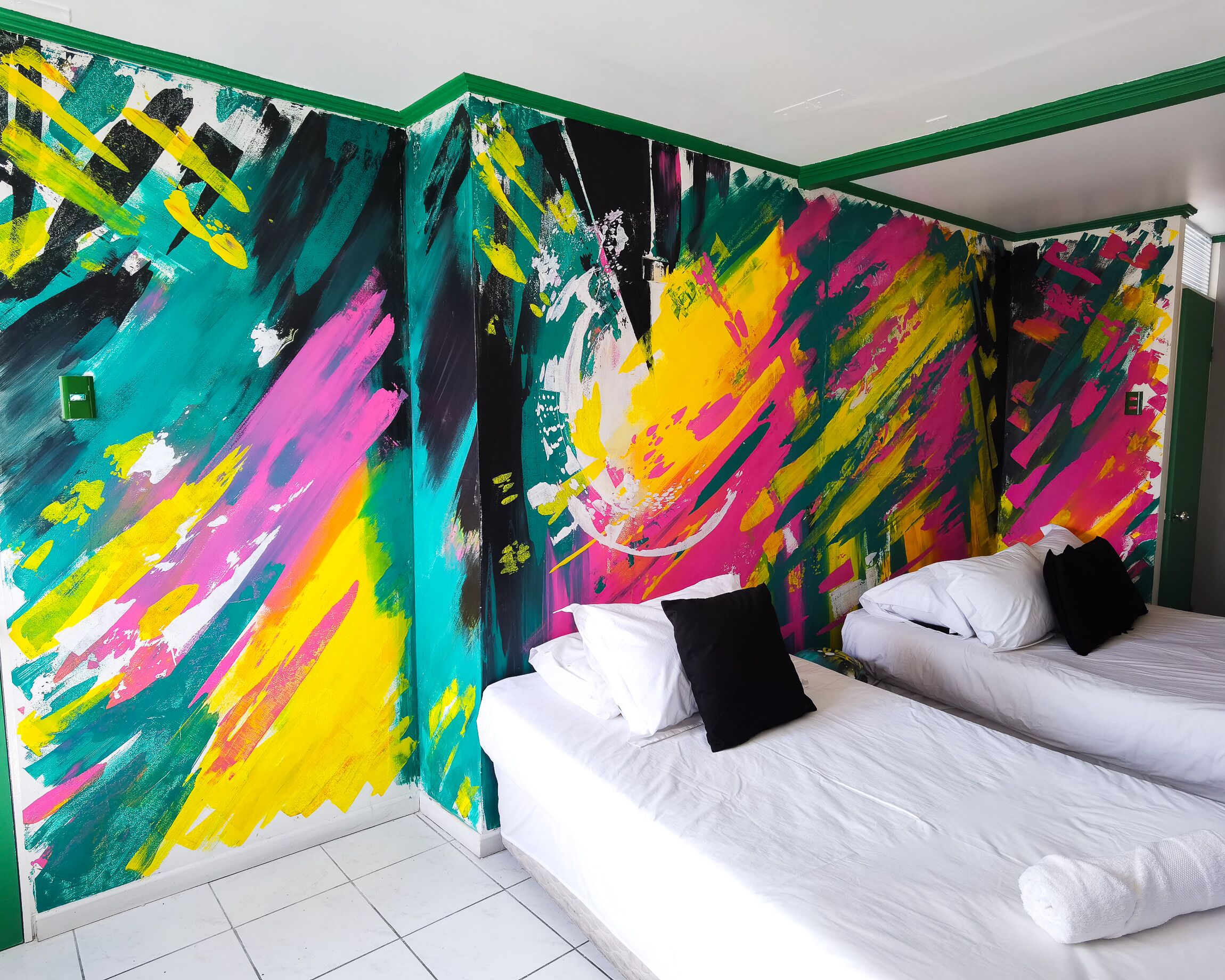 Una habitación con cama, ropa de cama blanca y un mural abstracto y colorido en la pared.