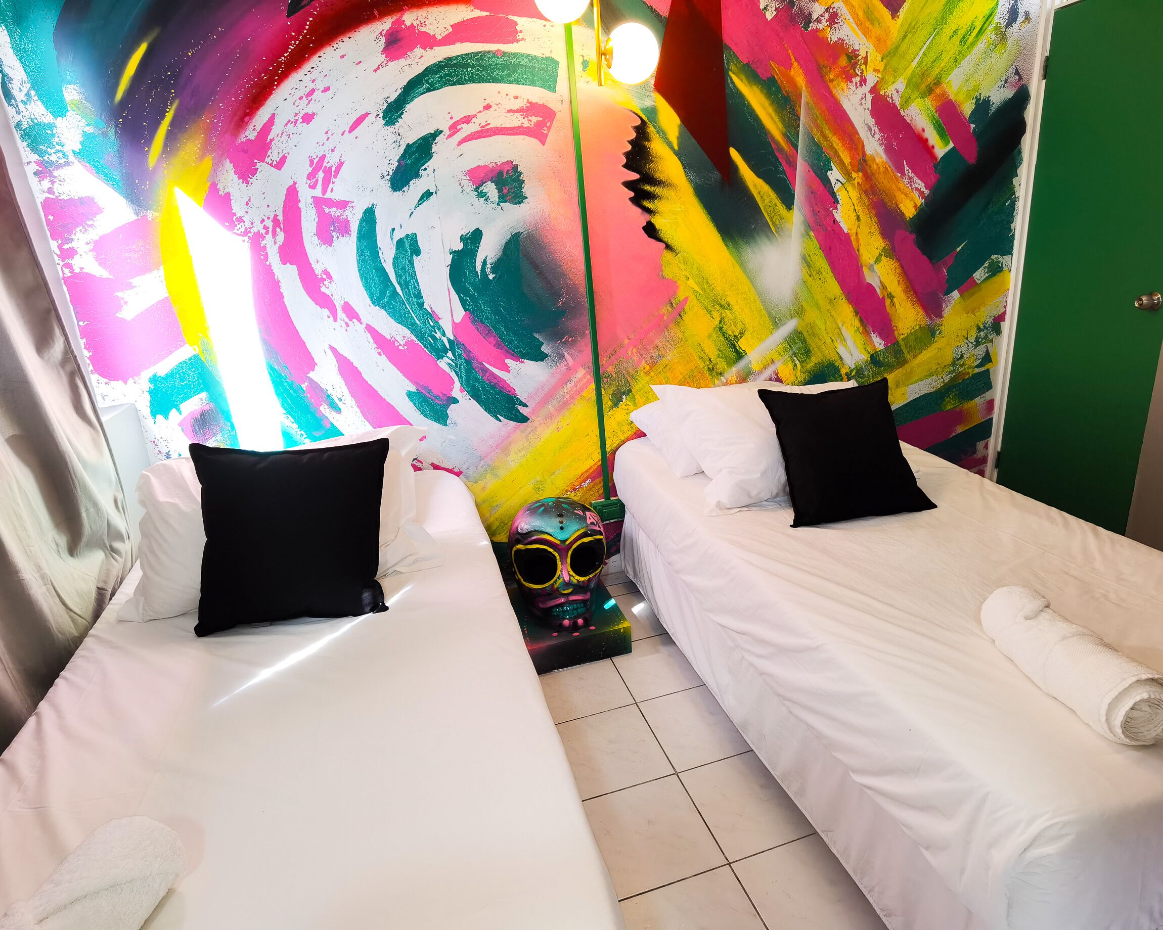 A room with two beds, each with white bedding and black pillows, and a colorful abstract mural on the wall behind them.