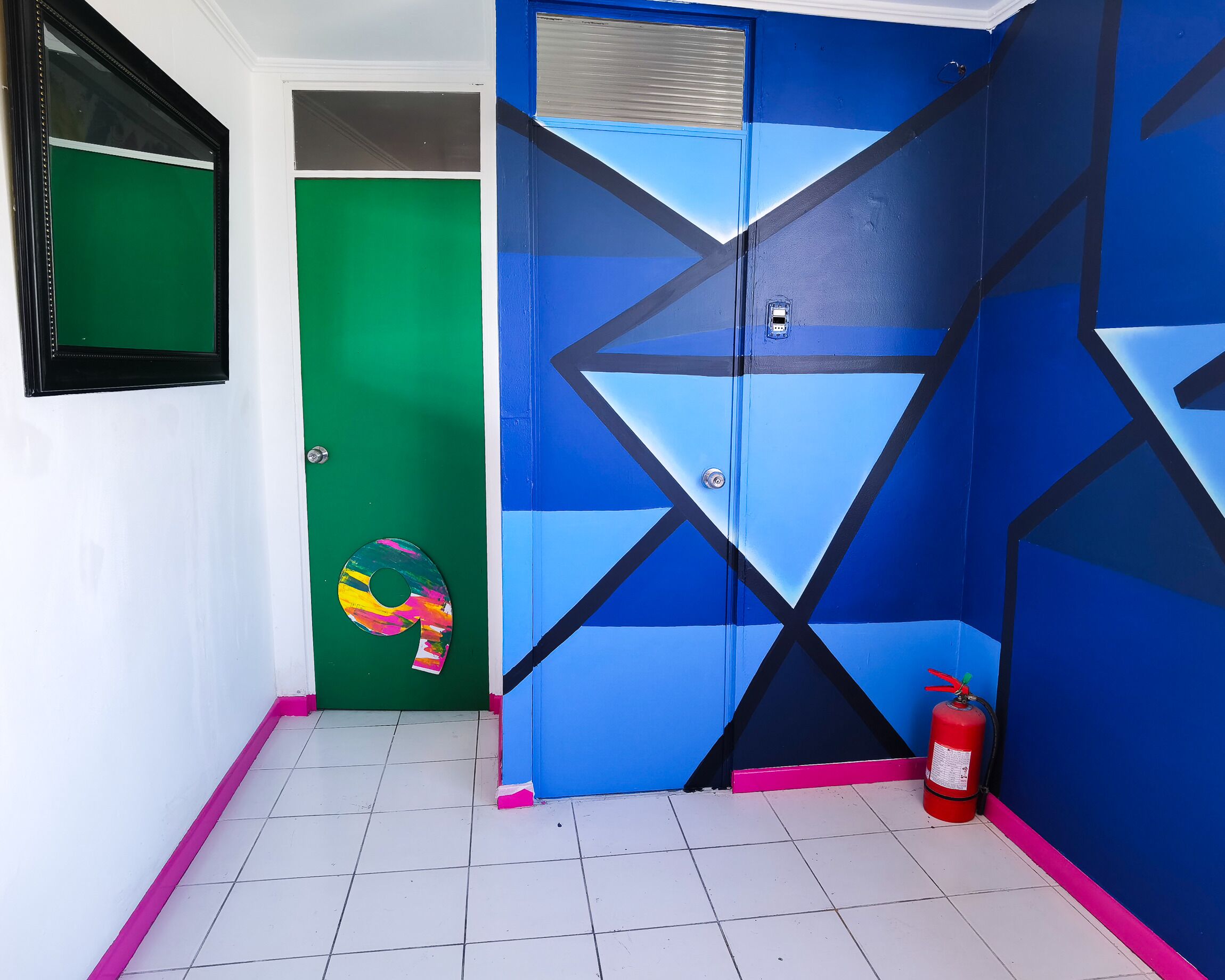Um corredor com uma porta verde, uma porta azul com padrões geométricos e um extintor no chão.