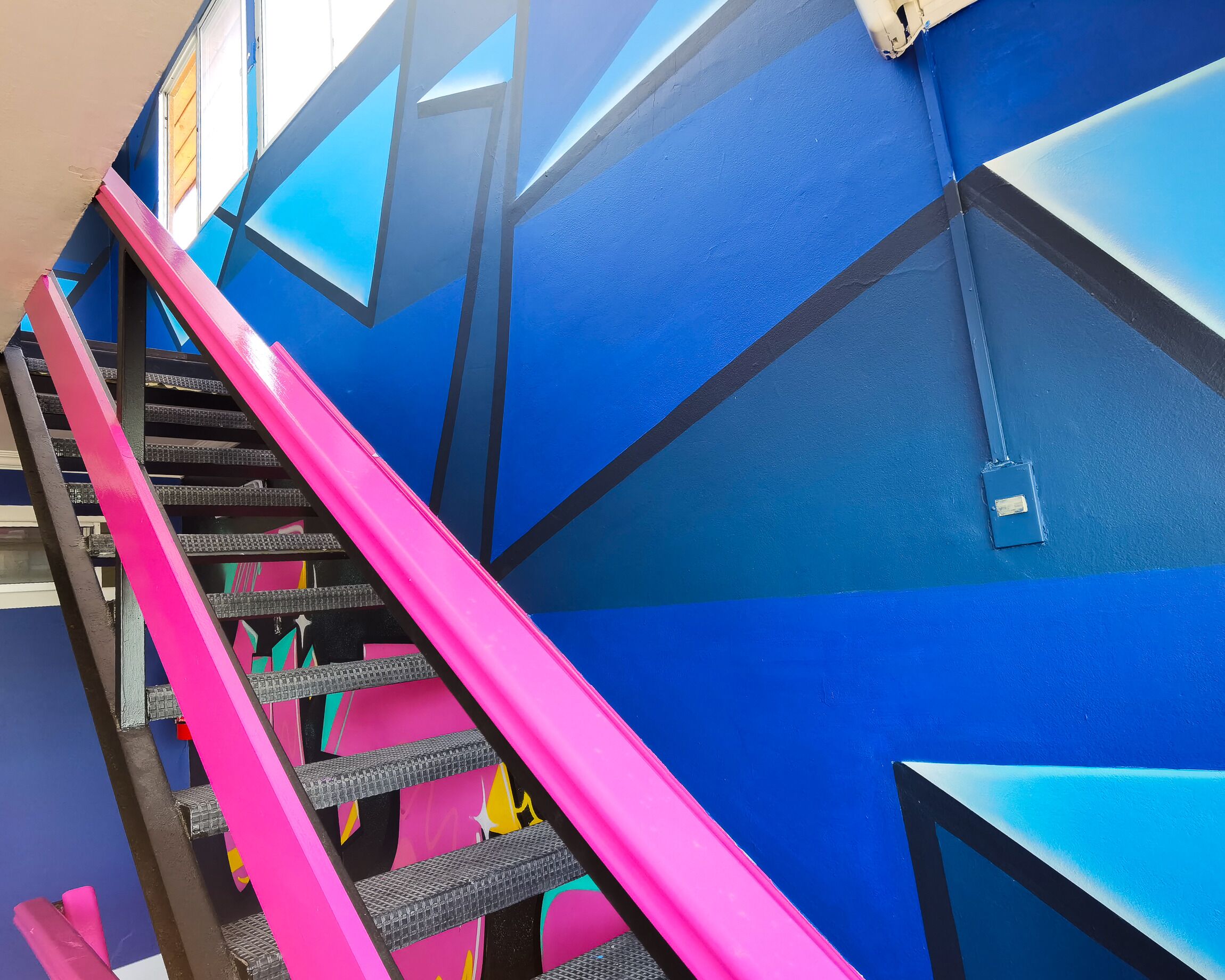 Una escalera con peldaños rosa y negros que conducen a una pared azul con patrones geométricos.