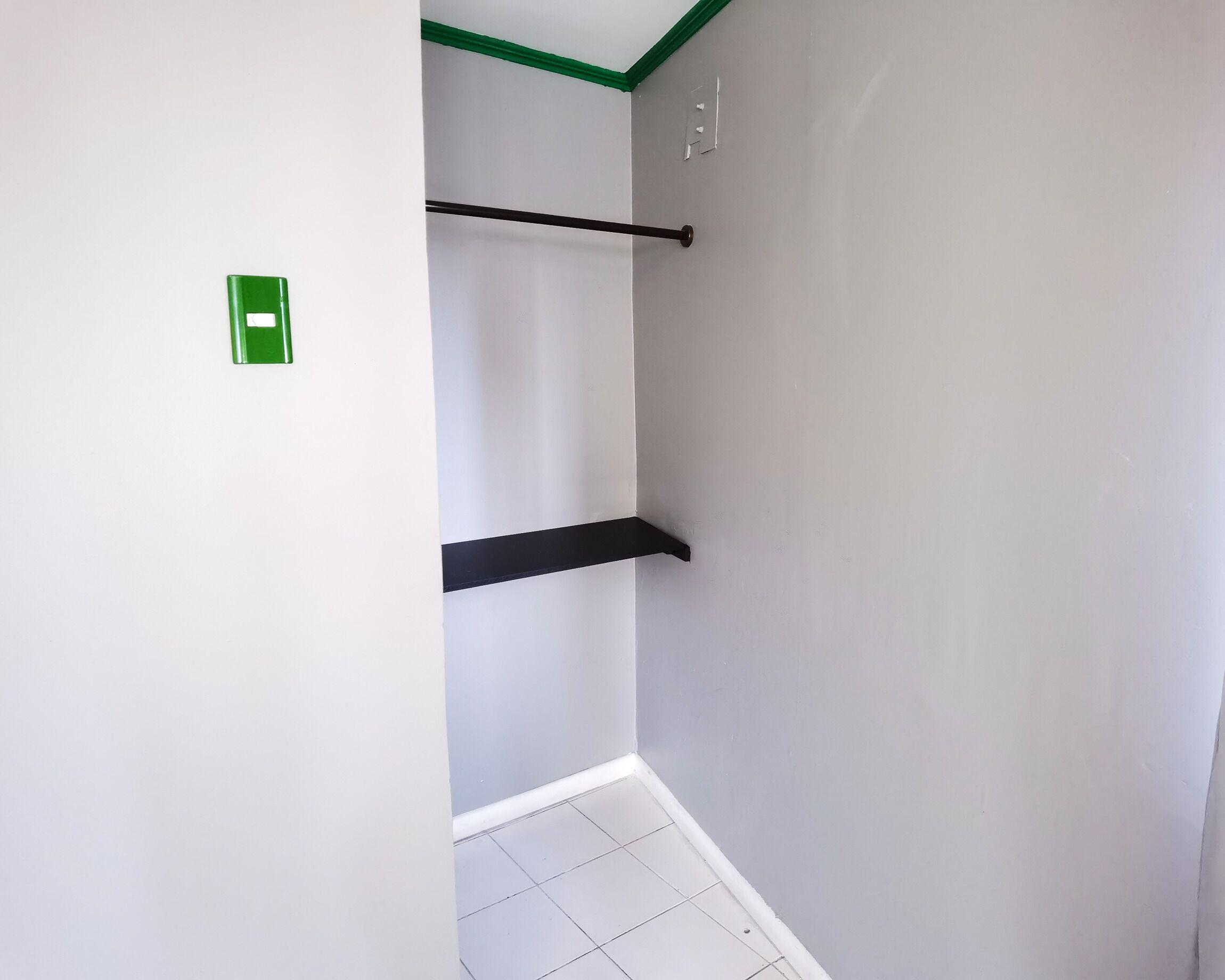 Ein schmaler Flur mit weißen Wänden und grüner Zierleiste an der Decke. An der Wand sind zwei schwarze Regale angebracht, und ein grünes Notausgangsschild ist auf der linken Seite sichtbar.