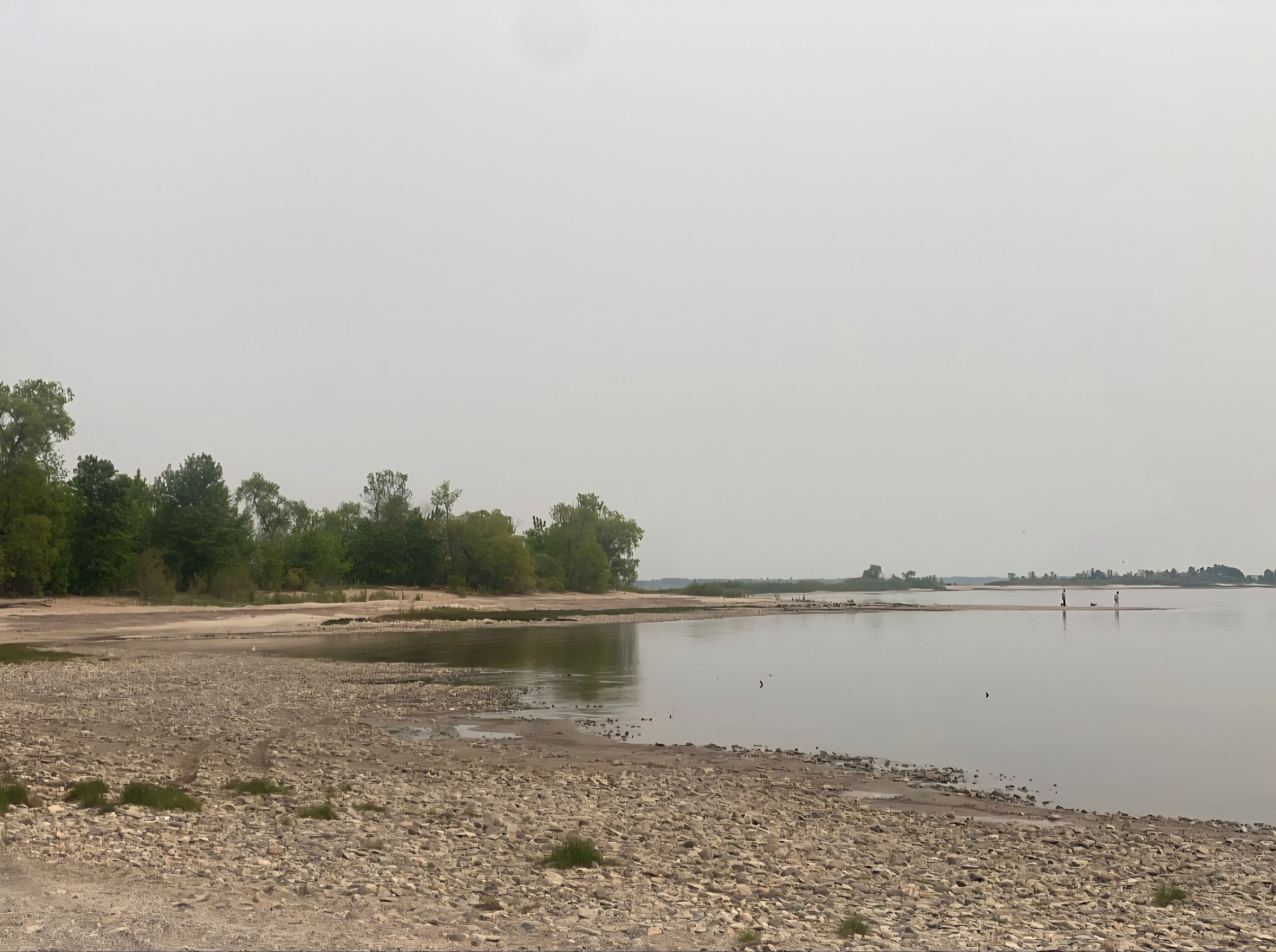 A rocky shoreline with a calm body of water, trees in the background, and a distant view of a small settlement.