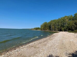 A clear view of a gravel beach with calm water, trees in the background, and a clear sky. - Family friendly/Cozy vibes (Beaconia)