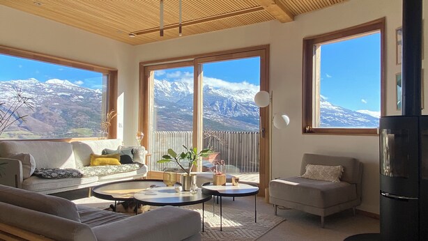 Ein gemütliches Wohnzimmer mit einer Couch, Sesseln und einem Couchtisch. Große Fenster bieten Blick auf die Berge.