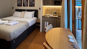 A compact hotel room with a bed, a small kitchenette, a dining table, and a view through a large window.