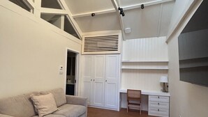 A modern living room with a sofa, a coffee table, and a desk with a chair. There is a wardrobe and a window with blinds.