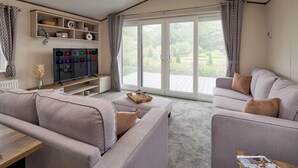 A modern living room with a large flat-screen TV, grey sofas, a wooden coffee table, and a sliding glass door leading to an outdoor area.