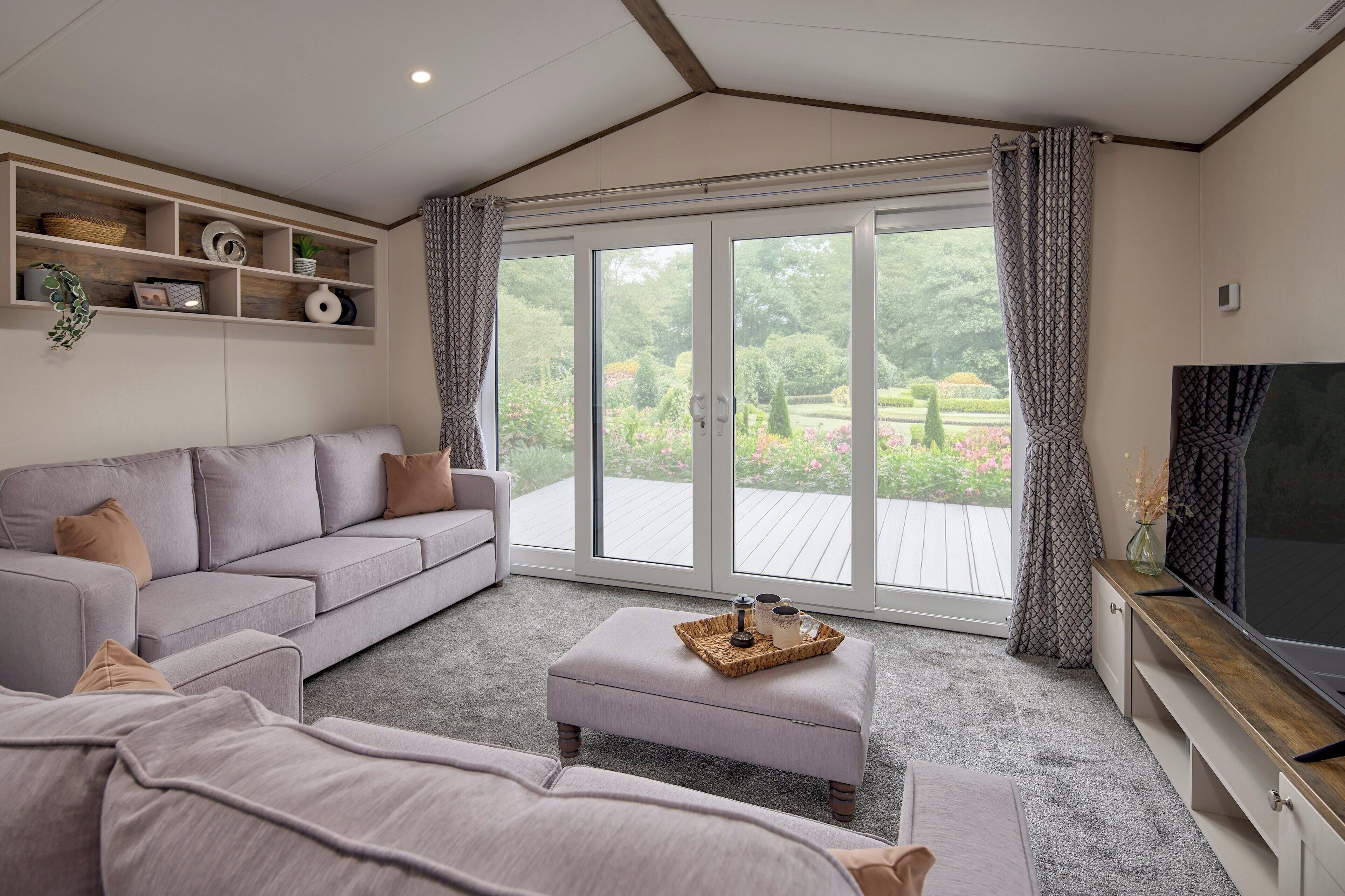 A modern living room with a grey sofa, a coffee table with decorative items, a flat-screen TV, and a large sliding glass door leading to an outdoor deck.