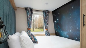 Un dormitorio con cama, almohadas blancas, cortinas estampadas, vistas a un parque y un mural de estrellas en la pared.