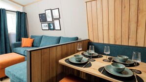 A dining area with a wooden table set for two, a blue sofa with orange cushions, and framed artwork on the wall. - Azure Horizons - Brook Glamping Pod Pet GV19 (Northampton)