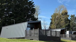 A modern building with a flat roof, a wooden deck, and a paved pathway leading to it. - Azure Horizons - Brook Glamping Pod Pet GV19 (Northampton)