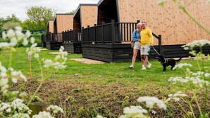 A row of wooden cabins with black railings, a grassy area in front, and a person walking a dog. - Azure Horizons - Brook Glamping Pod Pet GV19 (Northampton)