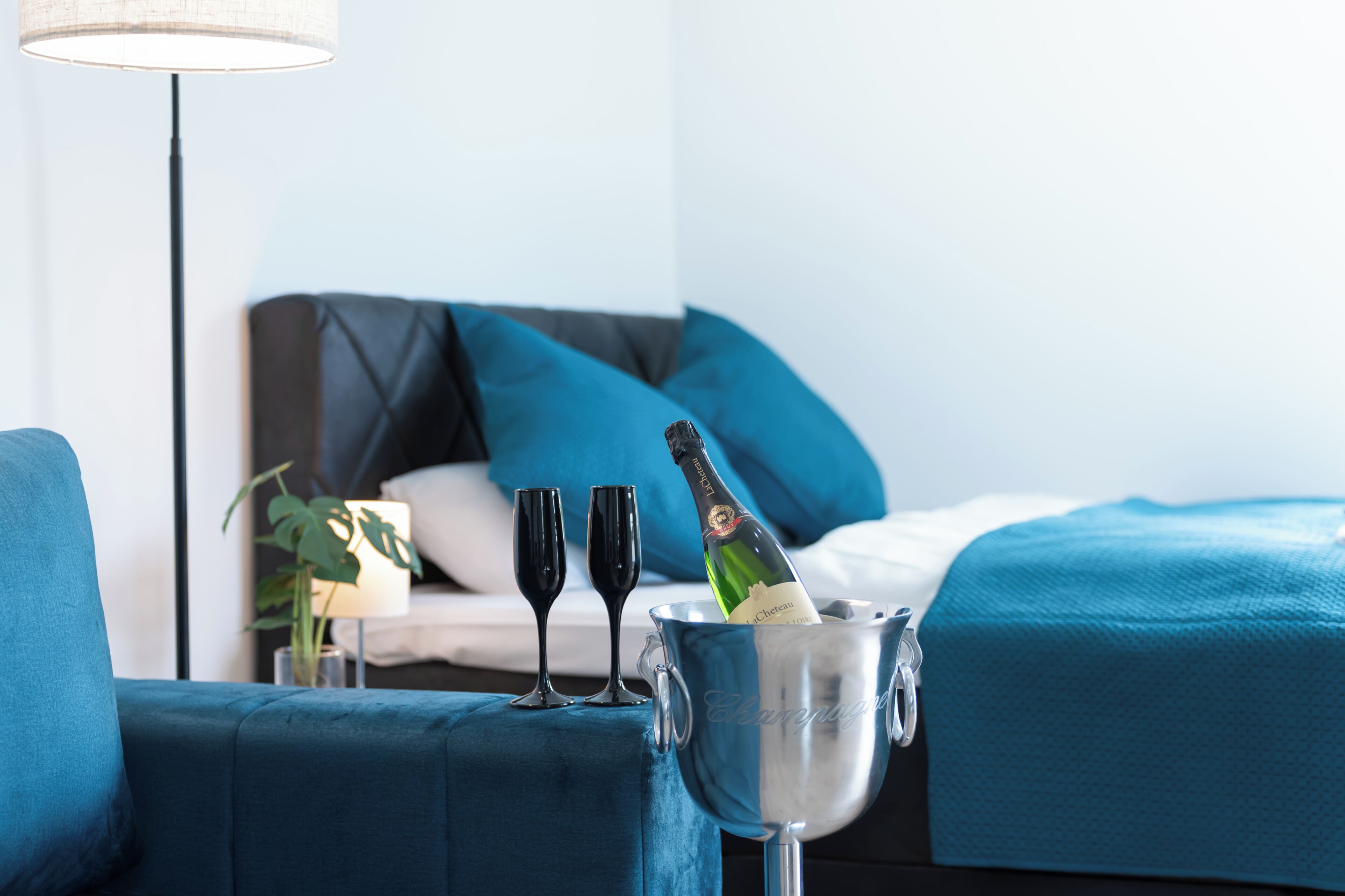 Une chambre d’hôtel avec un lit, un fauteuil bleu rembourré, une table de chevet avec une lampe, et un seau contenant deux verres et une bouteille de champagne.