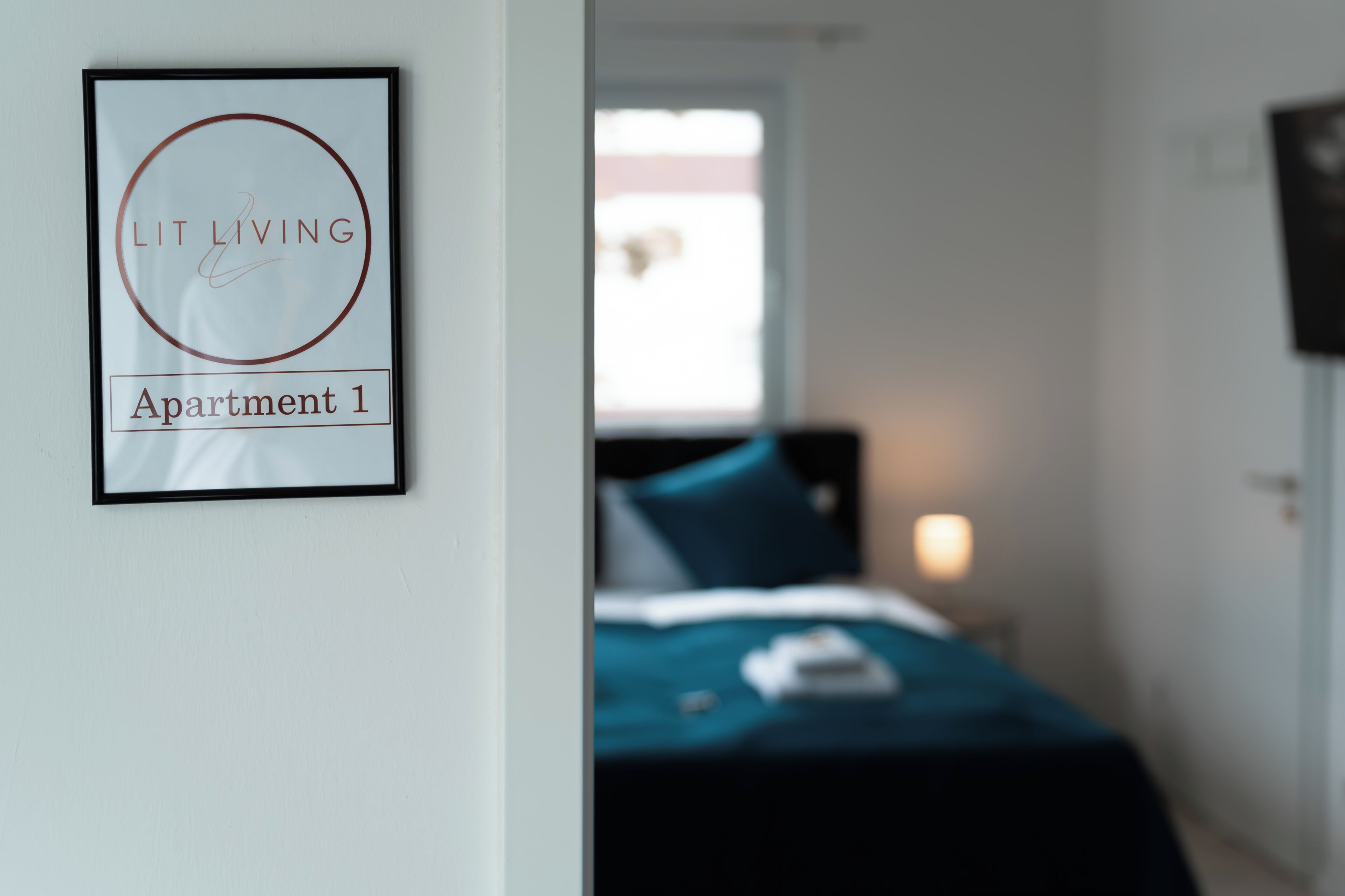 A framed sign on a wall, a bed with a blue cover, and a window with daylight.