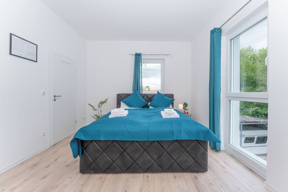 A bedroom with a large bed, a window with curtains, a door, and a framed notice on the wall.