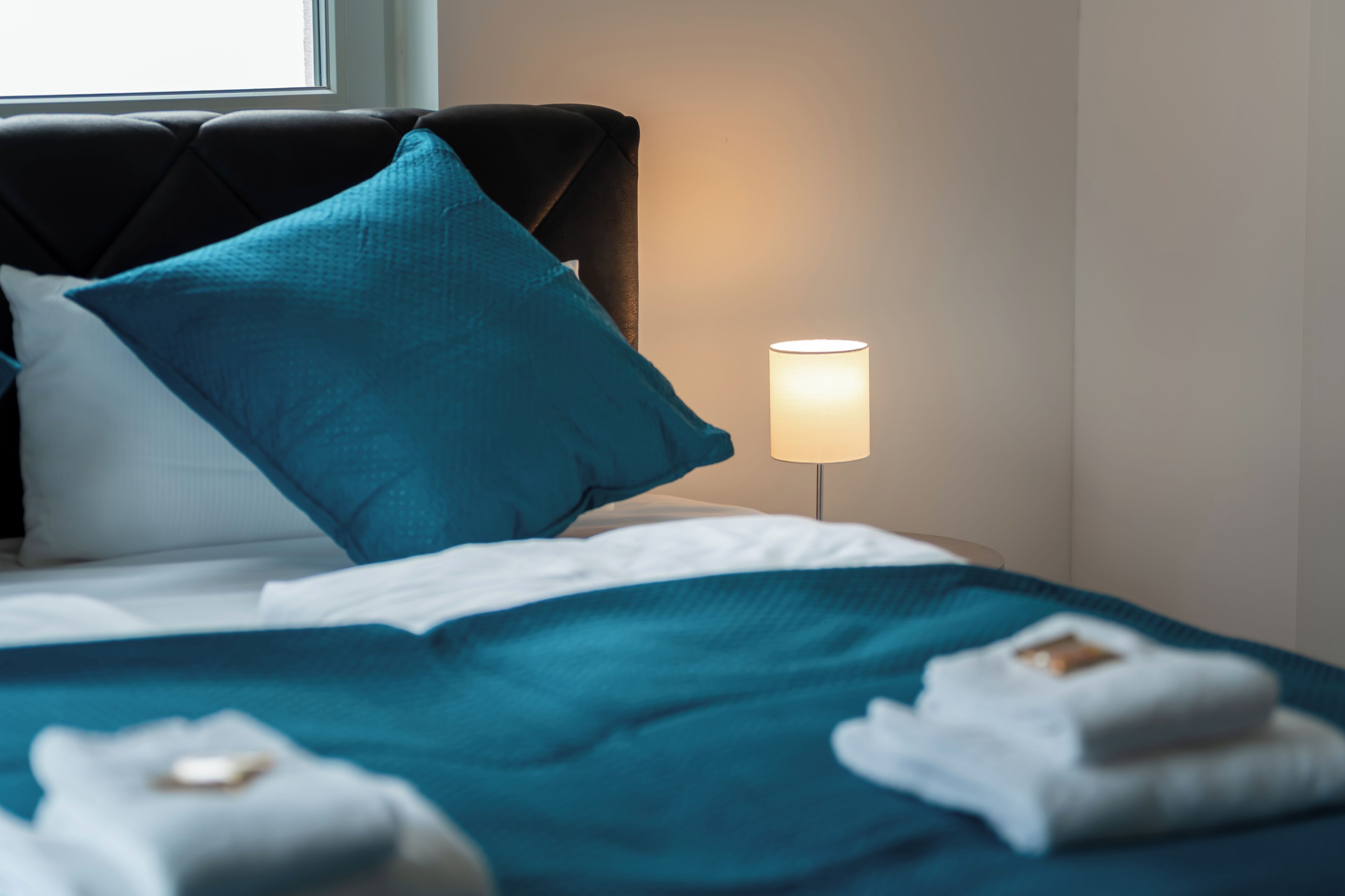 Ein ordentlich bezogenes Bett mit einem türkisfarbenen Überwurf, einem weißen Kopfkissen und einem dunklen Kopfteil. Eine Nachttischlampe ist eingeschaltet und beleuchtet den Raum.