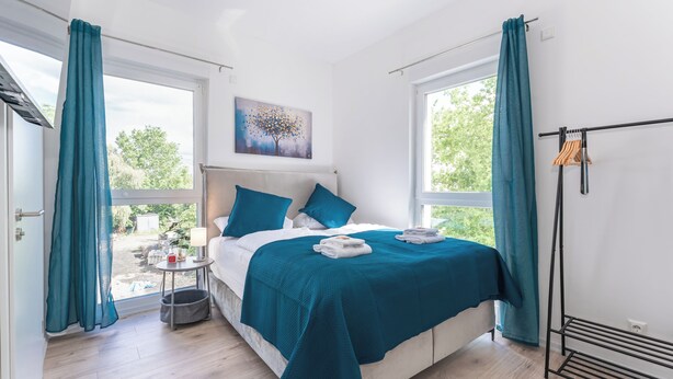 A modern bedroom with a bed, teal curtains, a window, and a painting. - Lit Living-Luxury-Box Spring - Parking (Altenstadt)