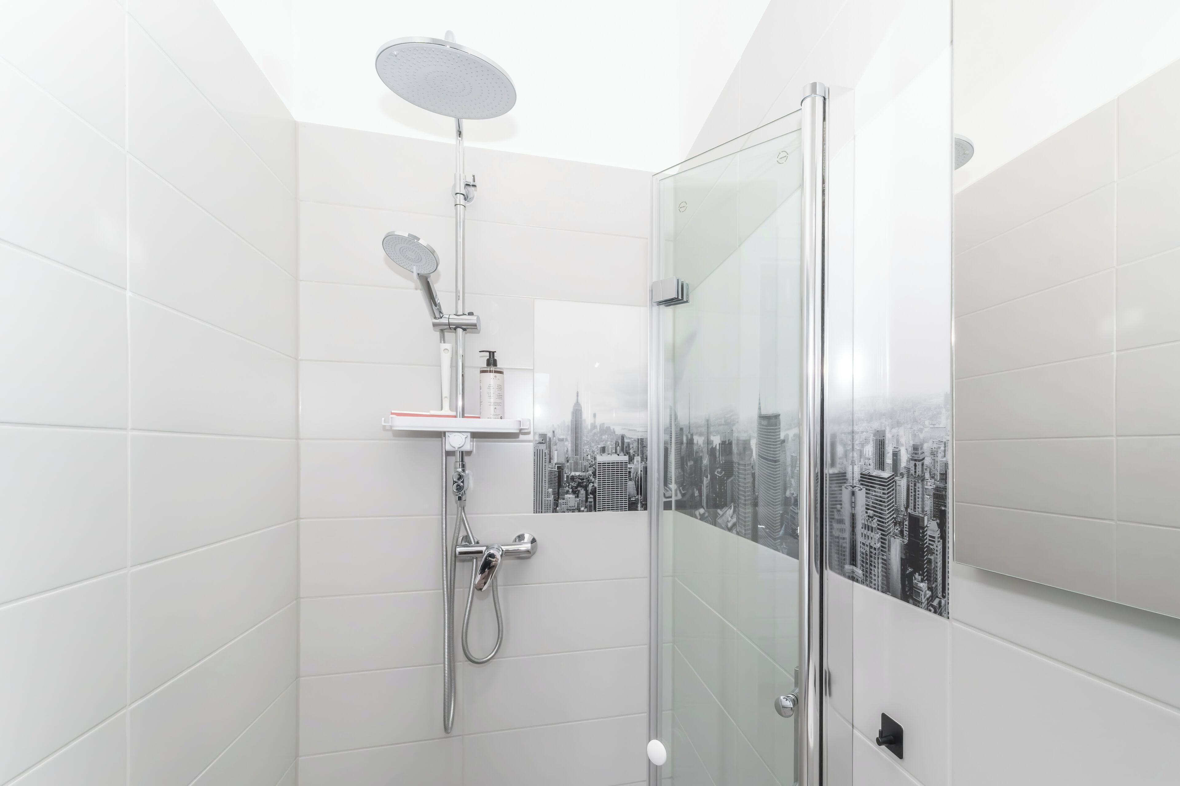 A modern bathroom with a glass shower enclosure, white tiled walls, and a cityscape view.