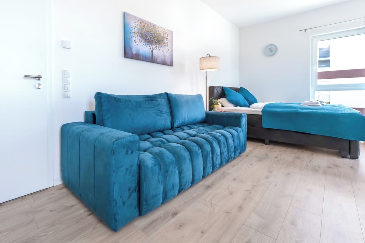 A modern bedroom with a blue sofa, a bed with matching blue bedding, a lamp, and a painting on the wall.
