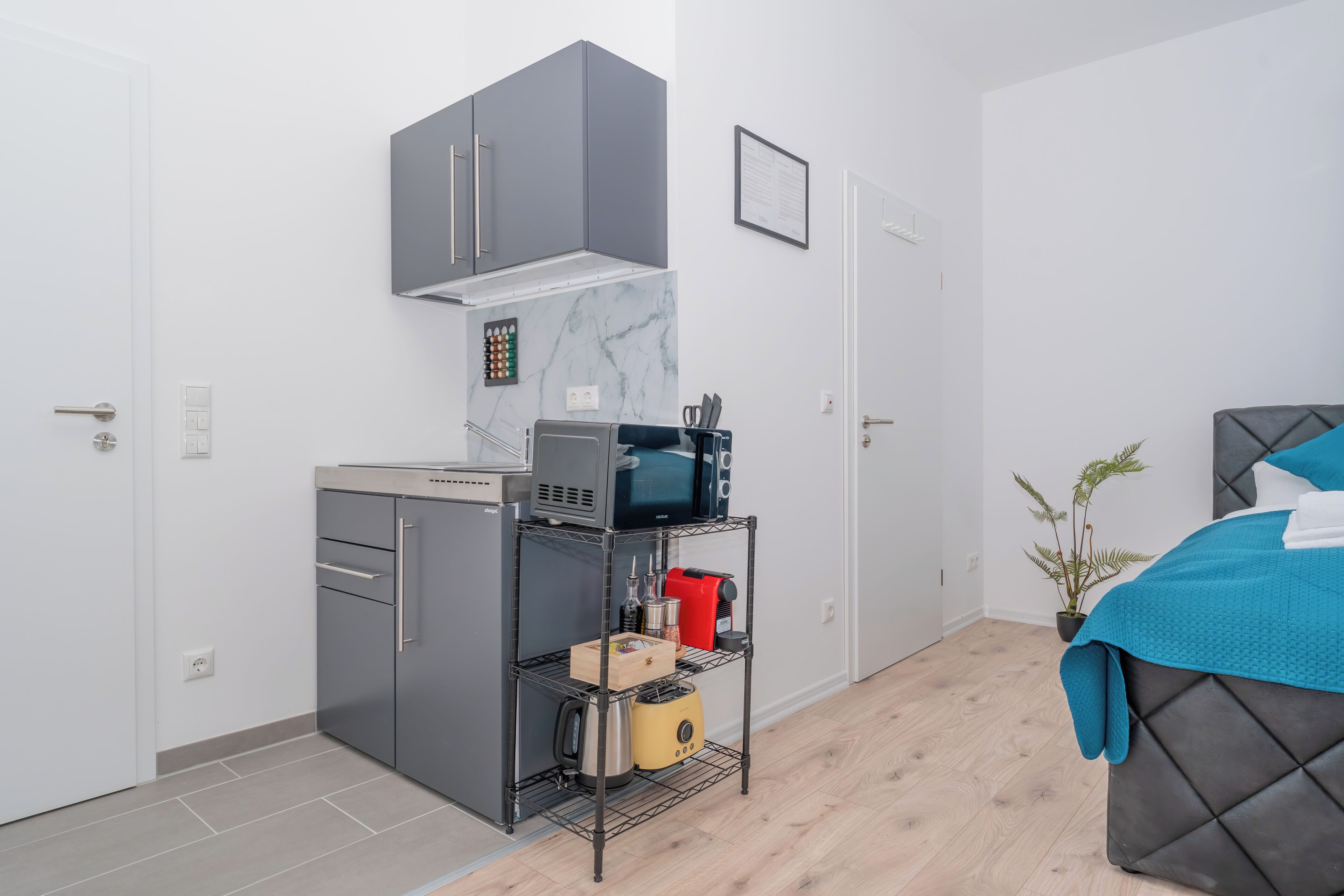 A compact room with a kitchenette, a bed with a blue blanket, and a potted plant.