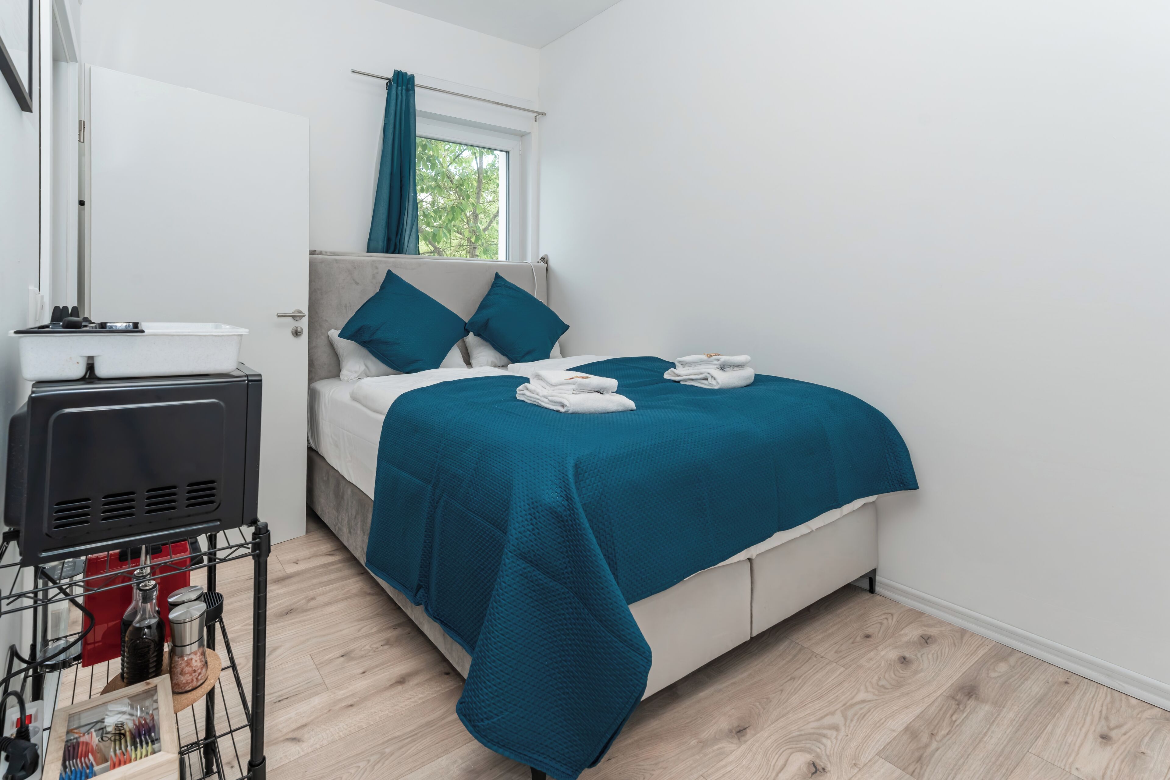 A modern hotel room with a bed, a window with curtains, a bedside table with a lamp, and a small kitchenette area.