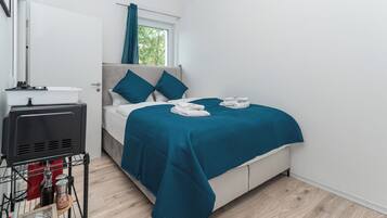 A modern hotel room with a bed, a window with curtains, a bedside table with a lamp, and a small kitchenette area.