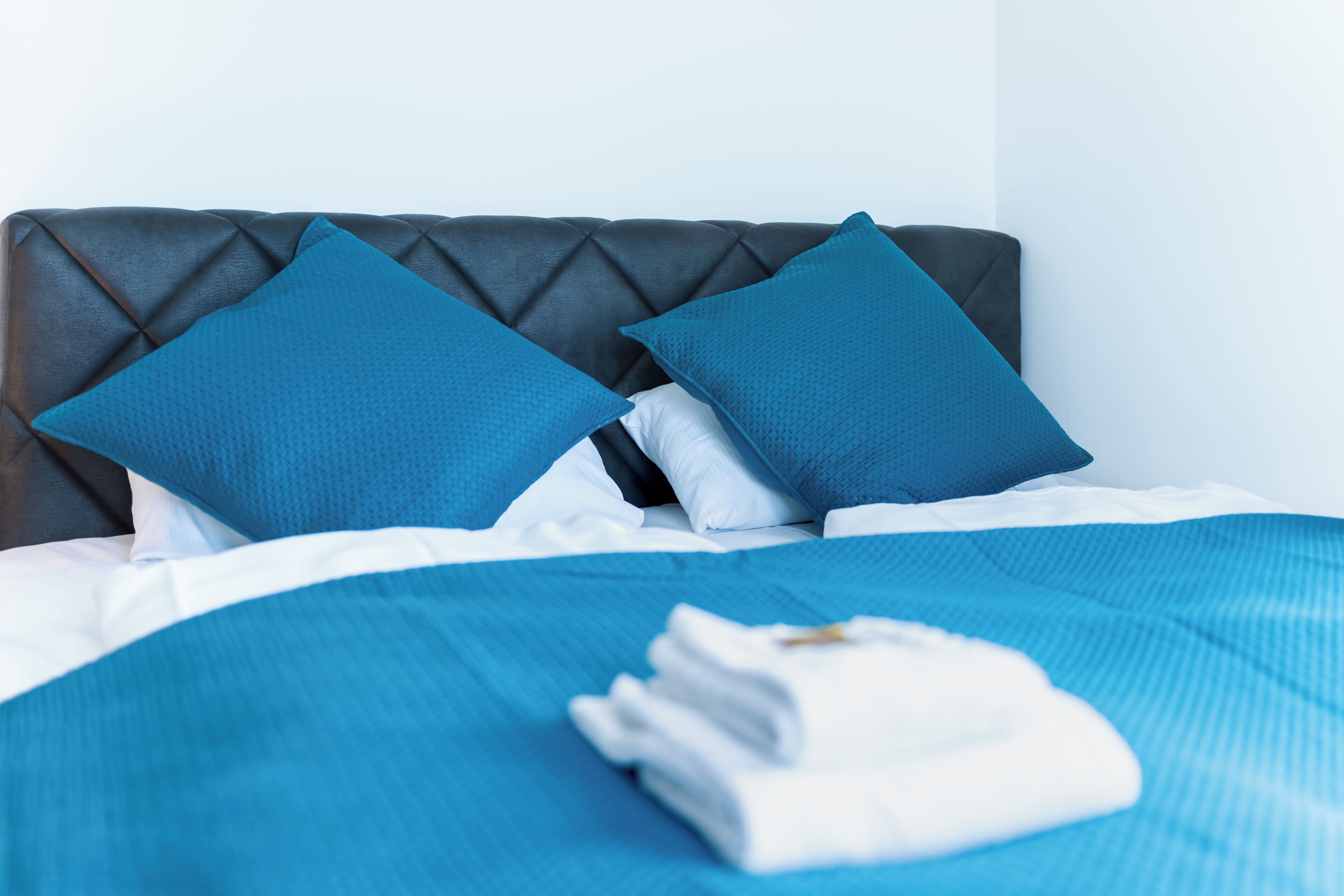 A neatly made bed with blue bedding and pillows, a folded white towel, and a dark headboard.