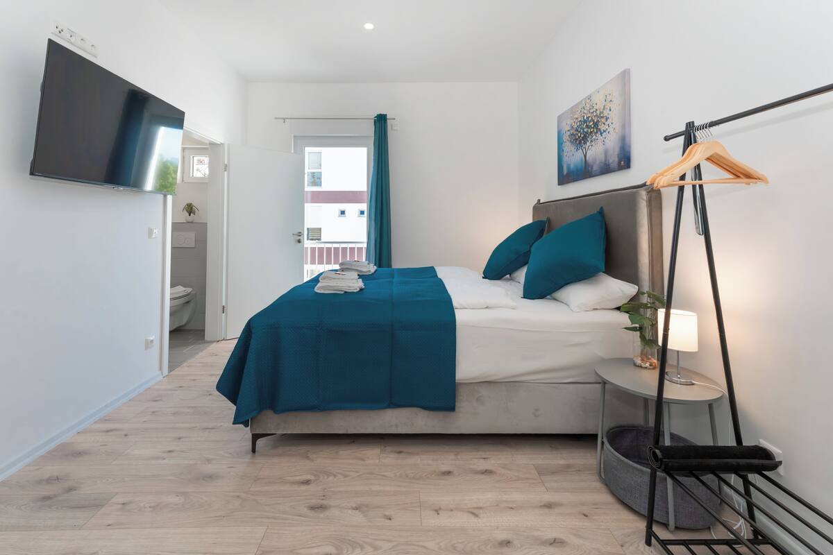 A modern bedroom with a bed, a TV mounted on the wall, a bedside table with a lamp, and a view of the outside balcony.