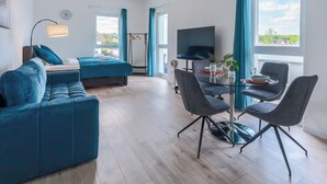 A modern living room with a blue sofa, a dining table with chairs, a TV, and a balcony with a view. - Lit Living-Luxury-Box Spring - Parking (Altenstadt)