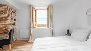 A bedroom with a brick wall, a bed with white bedding, a wooden chair, a nightstand with a lamp, and a window with curtains. - Le Duplex du Castel haut - Escapade Tarnaise (CASTELNAU DE MONTMIRAL)