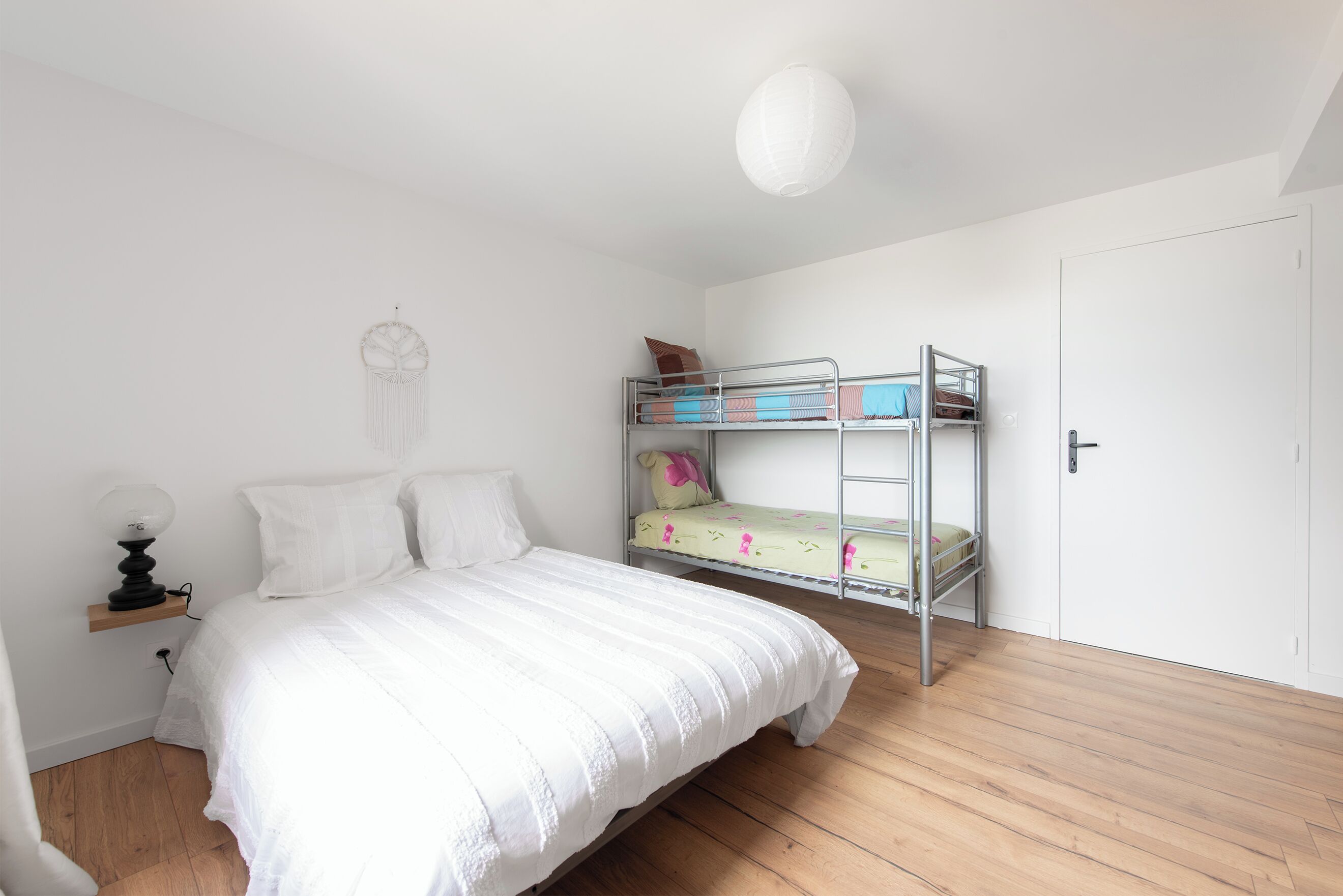 Ein Schlafzimmer mit Etagenbett, einem Einzelbett, einem Nachttisch mit Lampe und einem an der Wand hängenden Traumfänger.