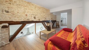 A loft bedroom with exposed wooden beams, a stone wall, a wooden floor, a bed with red bedding, and a small table with a basket. - Le Duplex du Castel haut - Escapade Tarnaise (CASTELNAU DE MONTMIRAL)