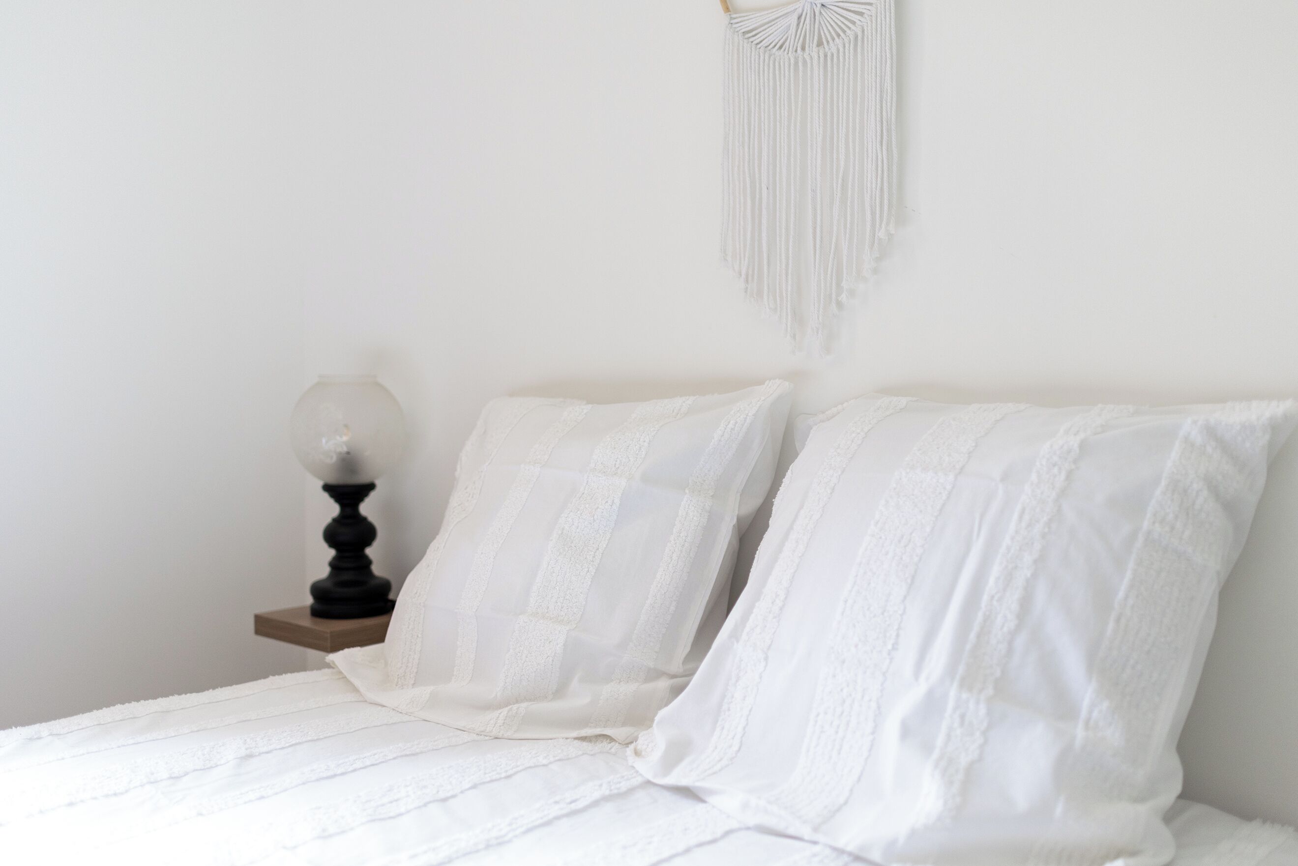 A bed with white bedding and a macrame wall hanging.