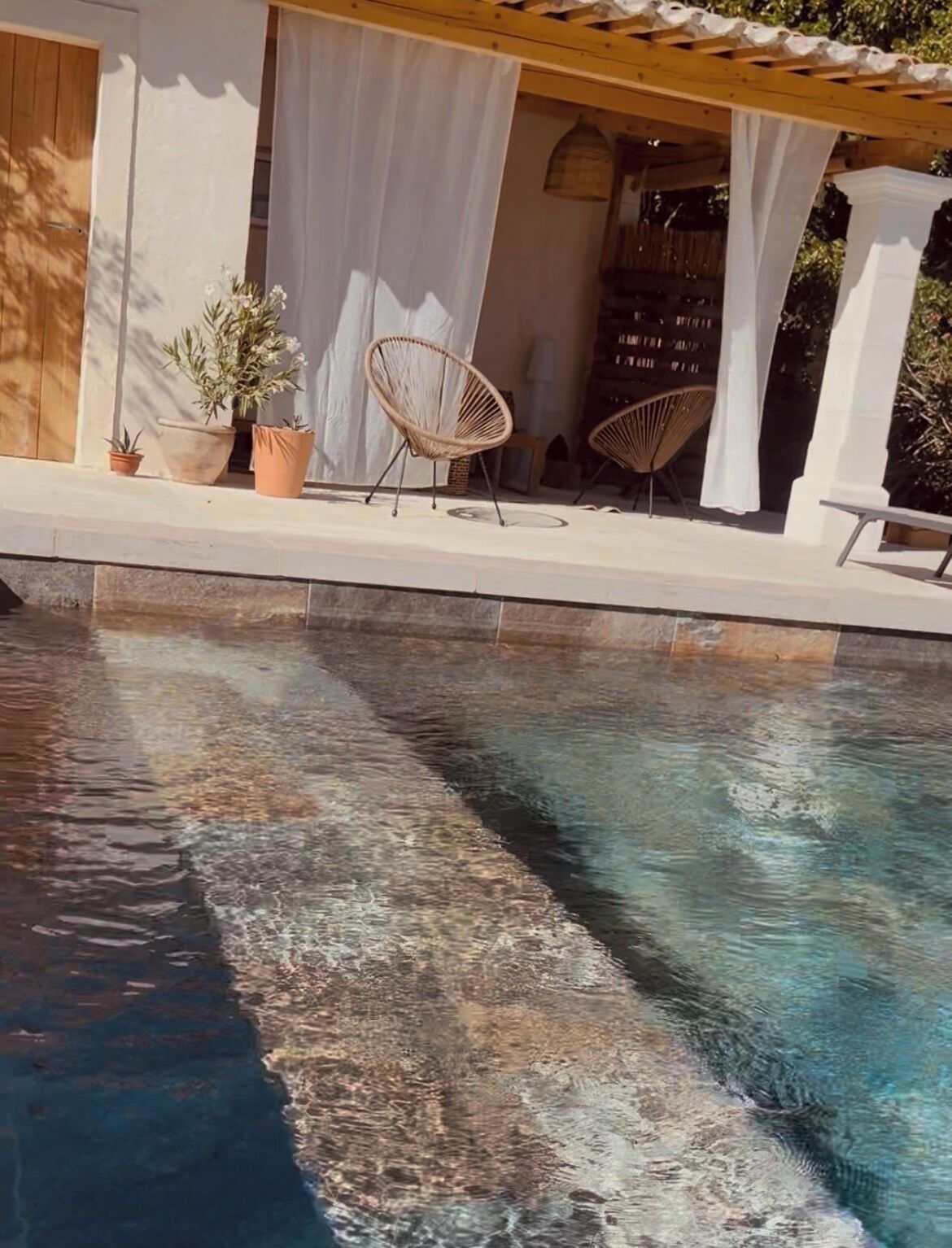 A swimming pool with clear water, surrounded by a covered patio area with chairs and potted plants.