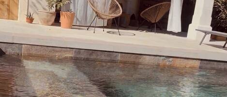 A swimming pool with clear water, surrounded by a covered patio area with chairs and potted plants.