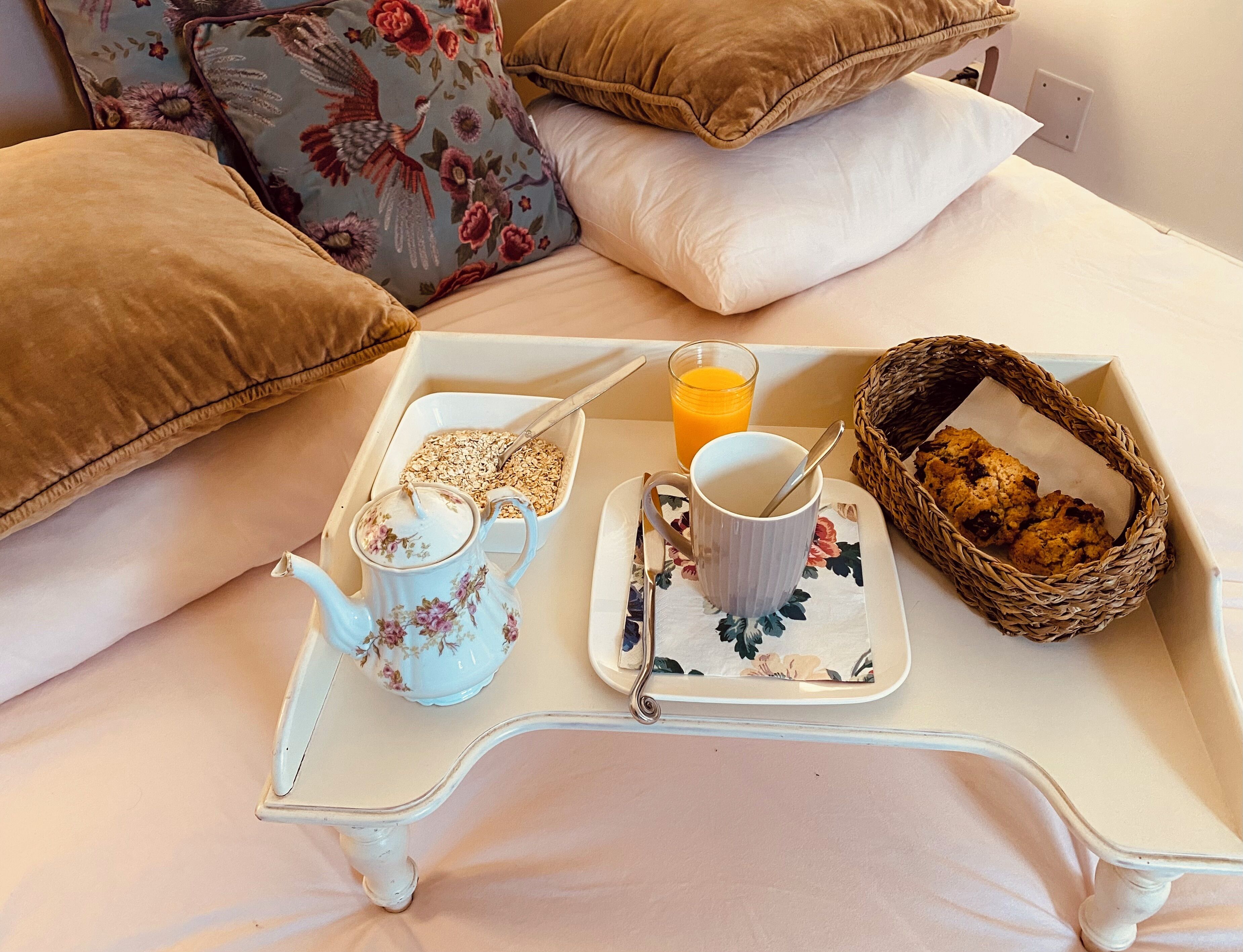 A breakfast tray on a bed with a teapot, mug, cereal, orange juice, and a basket of cookies.